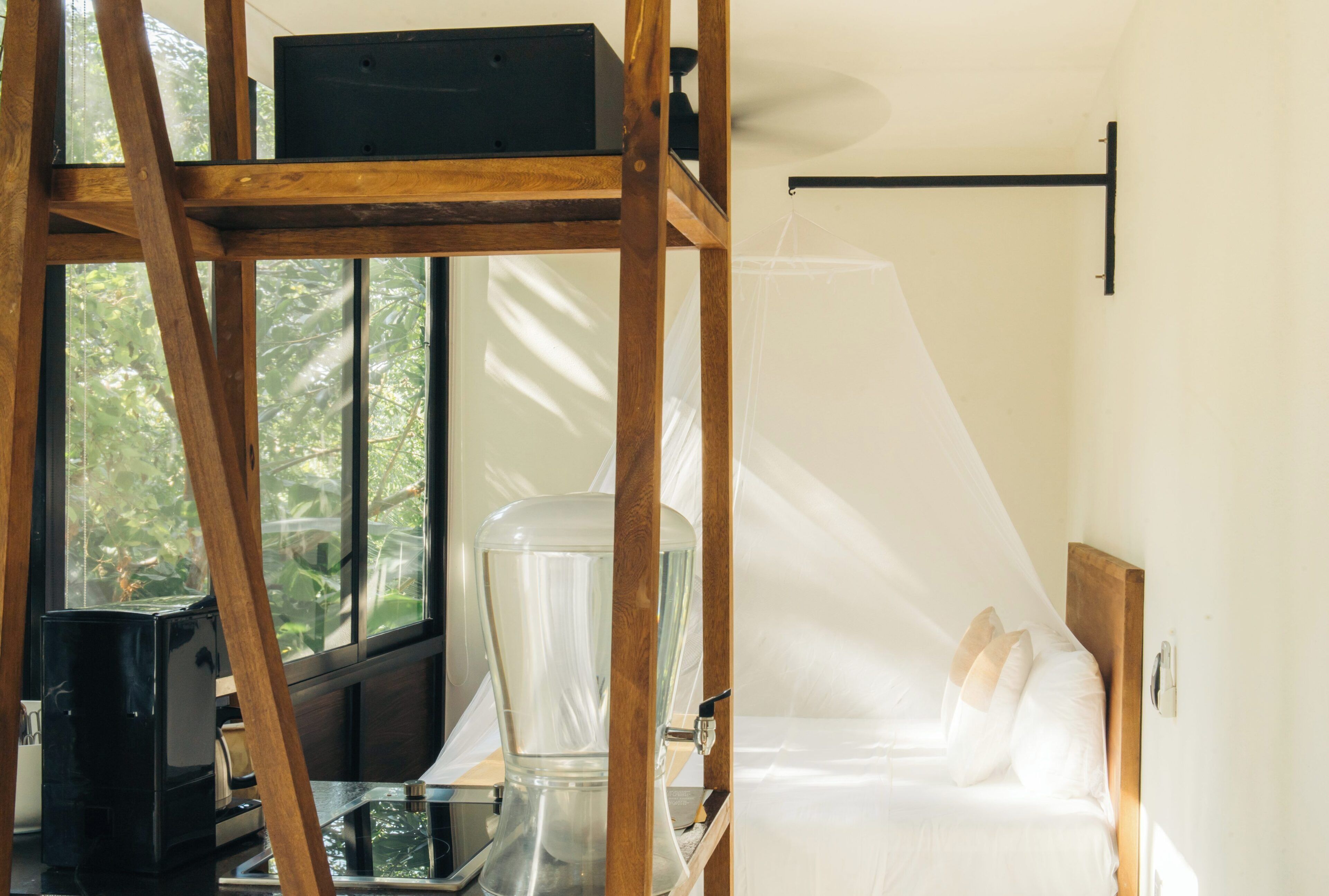 Una habitación con una cama, una escalera de madera, un dispensador de agua de vidrio y una ventana con vista a árboles.