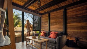 A room with a wooden floor, a sofa with red cushions, a wooden table, and a large window with a view of palm trees.