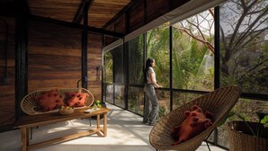 A room with a large window, a wooden bench, a wicker chair, and a table with fruits.