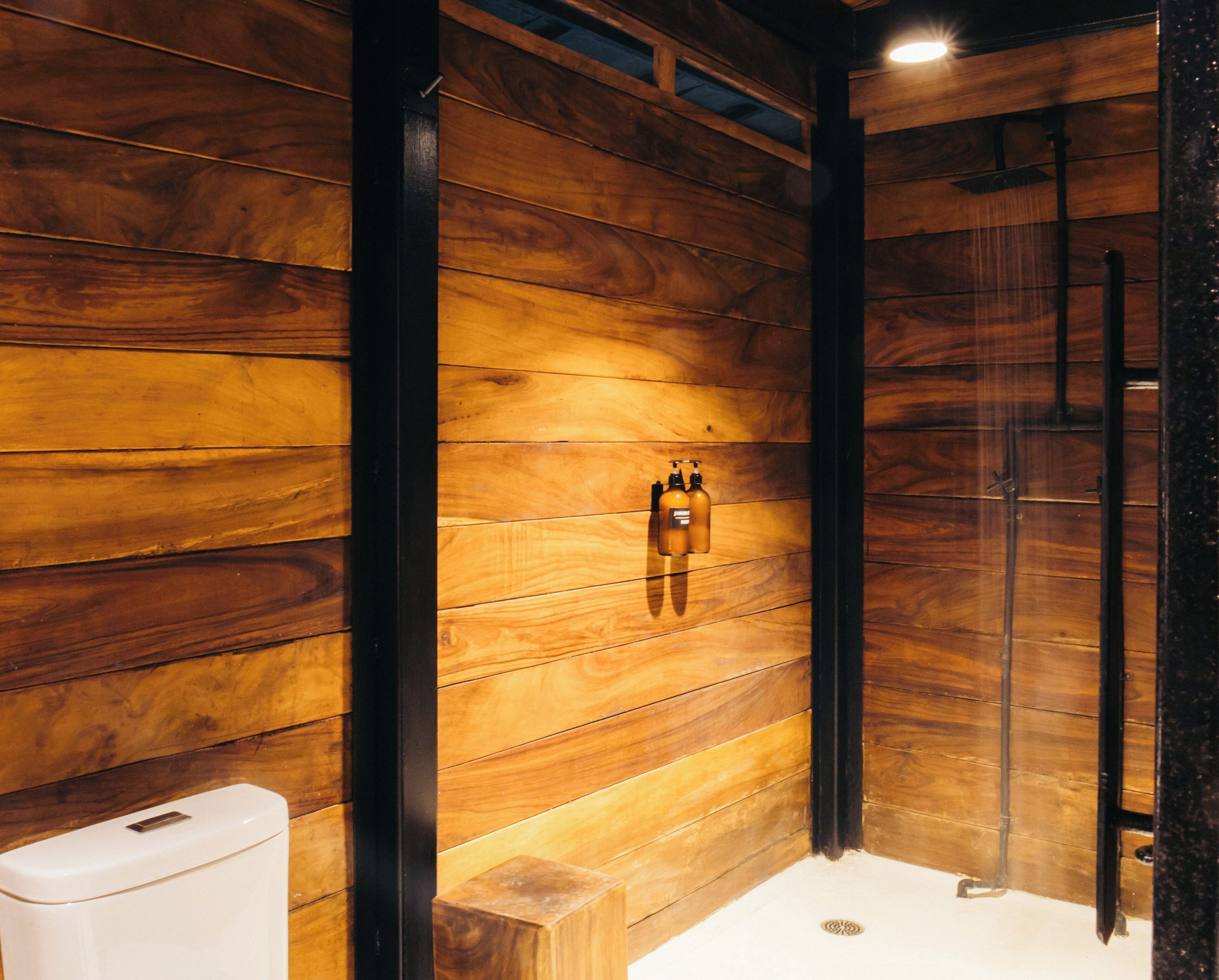 A wooden-paneled bathroom with a toilet, a shower, and two bottles of shampoo on the wall.