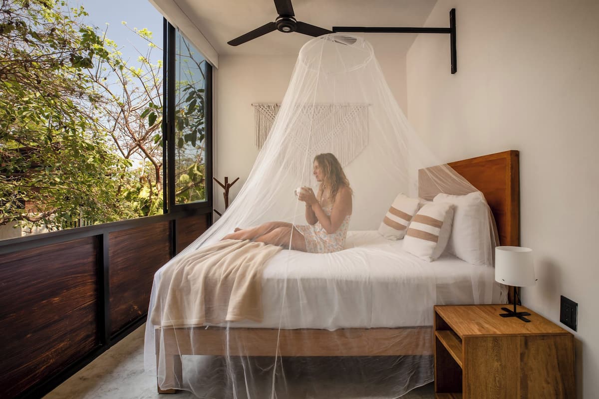 Un dormitorio con cama, mesita de noche, ventilador de techo y vista a árboles desde afuera.