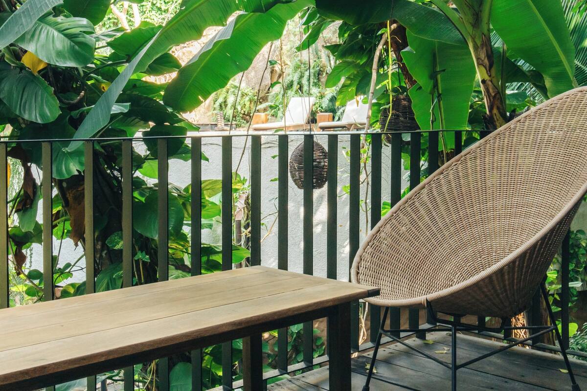 Una silla de mimbre y una mesa de madera en un balcón con vistas a un follaje verde exuberante.