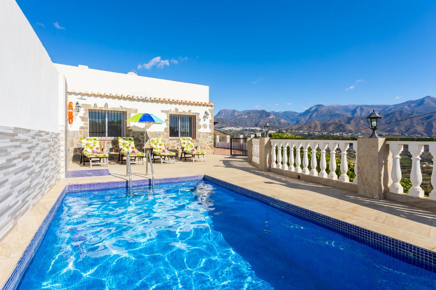 A swimming pool with a view of mountains, a white building with a balcony, and outdoor seating.