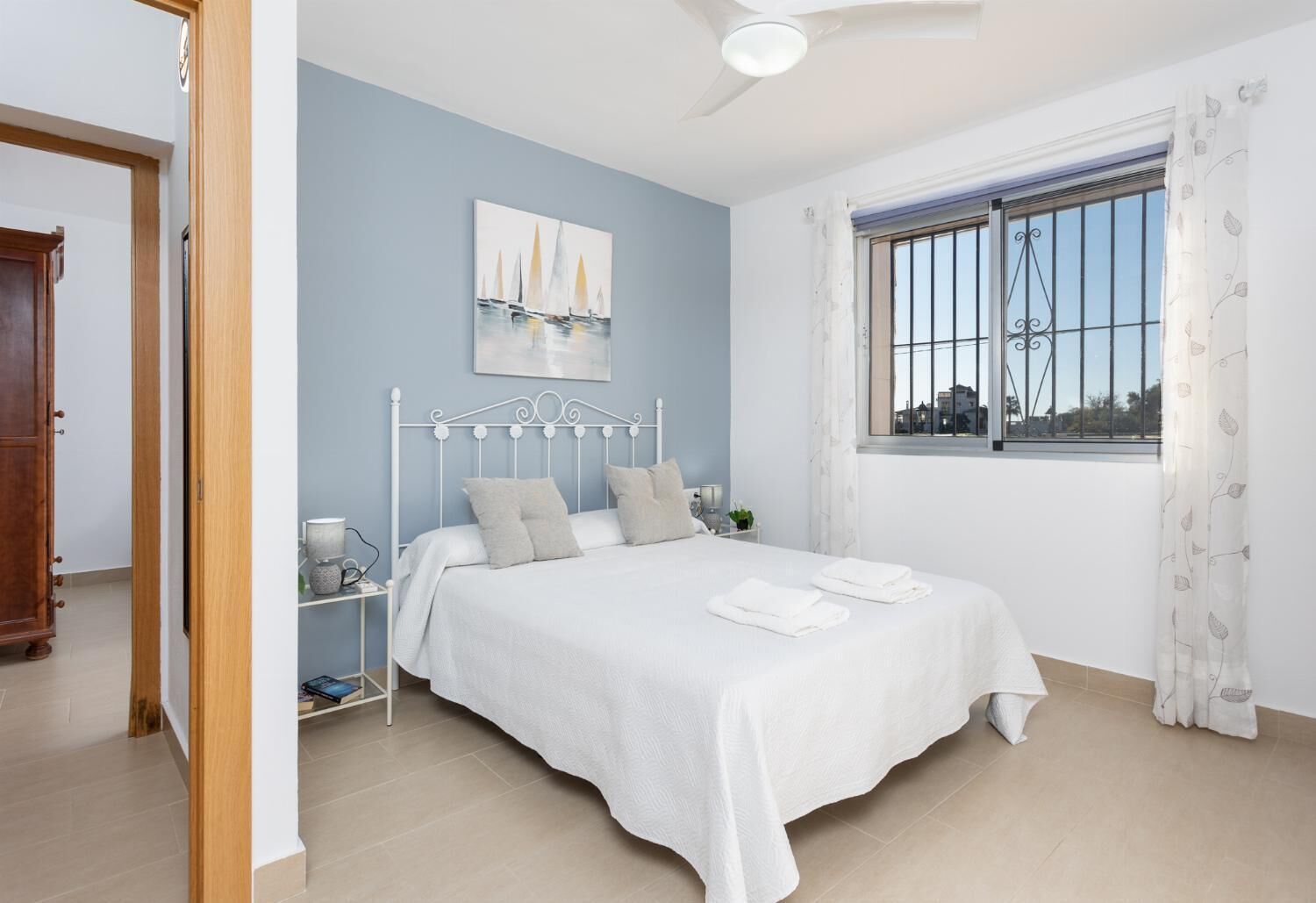 Un dormitorio con cama, mesita de noche, un cuadro, ventana con cortinas y vista a edificios.