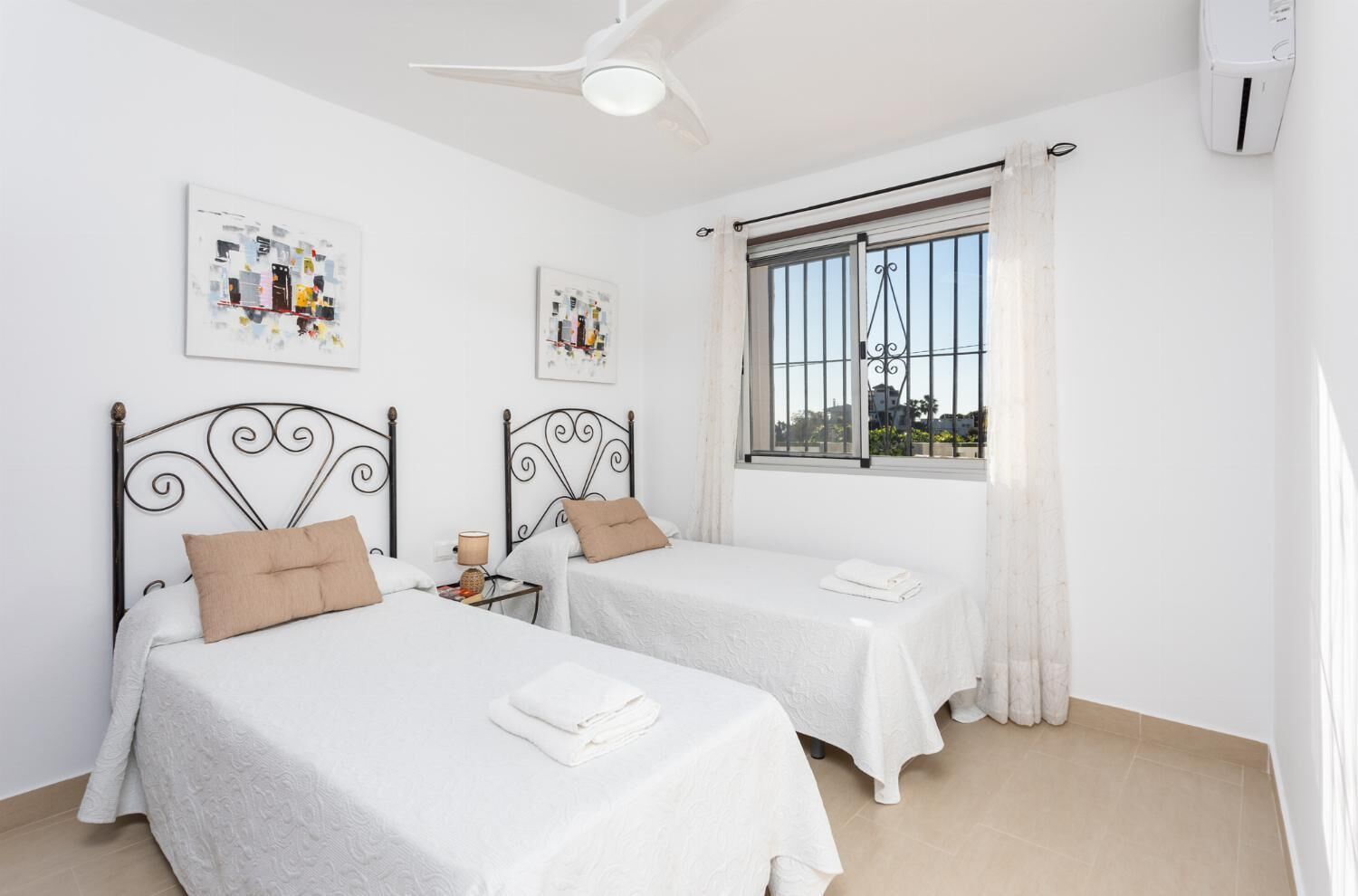 A bedroom with two beds, a ceiling fan, a window with bars, and artwork on the walls.