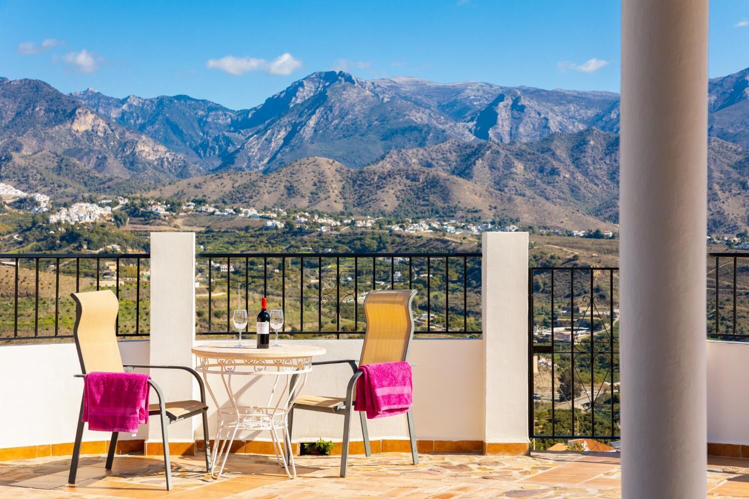 Un balcón con dos sillas, una mesa y una botella de vino. El balcón ofrece vistas a un paisaje montañoso con un pueblo al fondo.