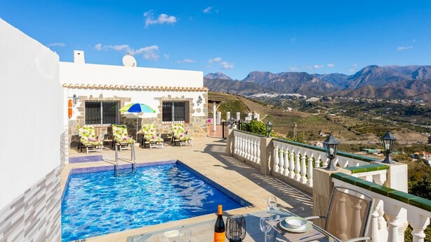 Una piscina al terrat amb vista despejada a les muntanyes, una zona de menjador amb taules i cadires, i un edifici blanc amb teulada enrajolada.