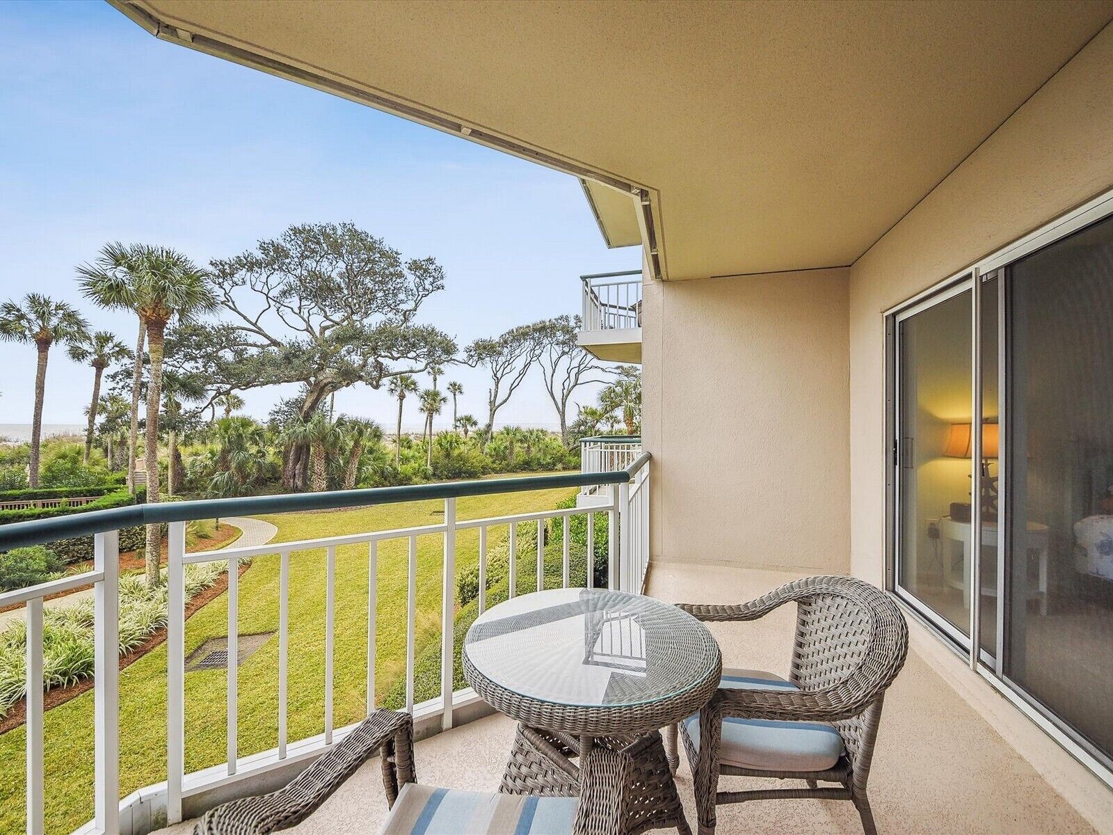 A balcony with wicker furniture, a glass table, and a view of a landscaped area with trees.
