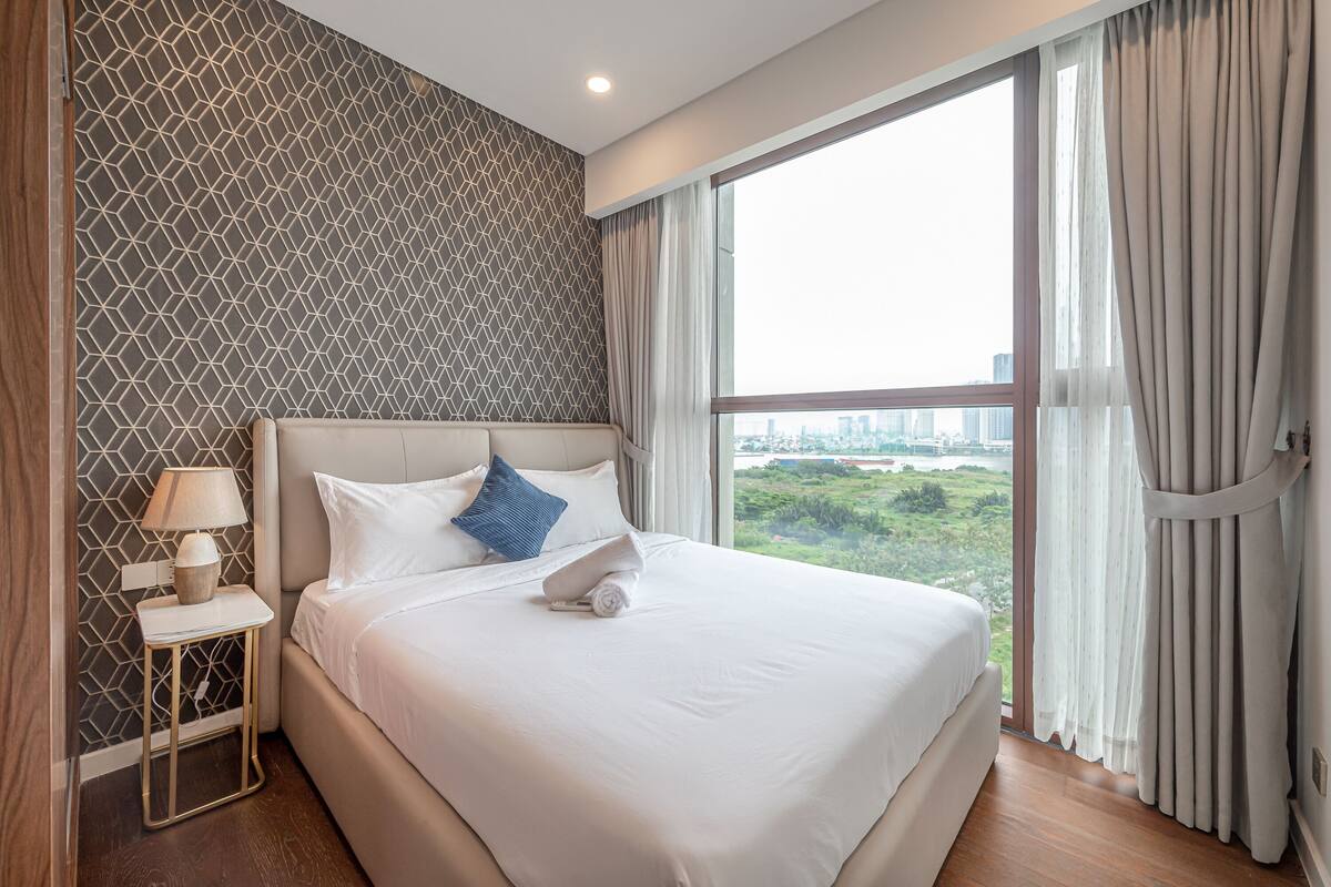 Habitación de hotel con una cama grande, una mesita de noche, una lámpara y vistas a un paisaje urbano a través de la ventana.