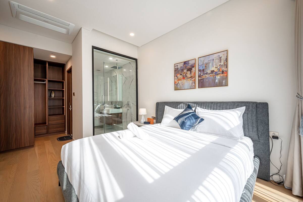 Una habitación de hotel moderna con una cama grande, un armario, ducha de vidrio y dos cuadros en la pared.