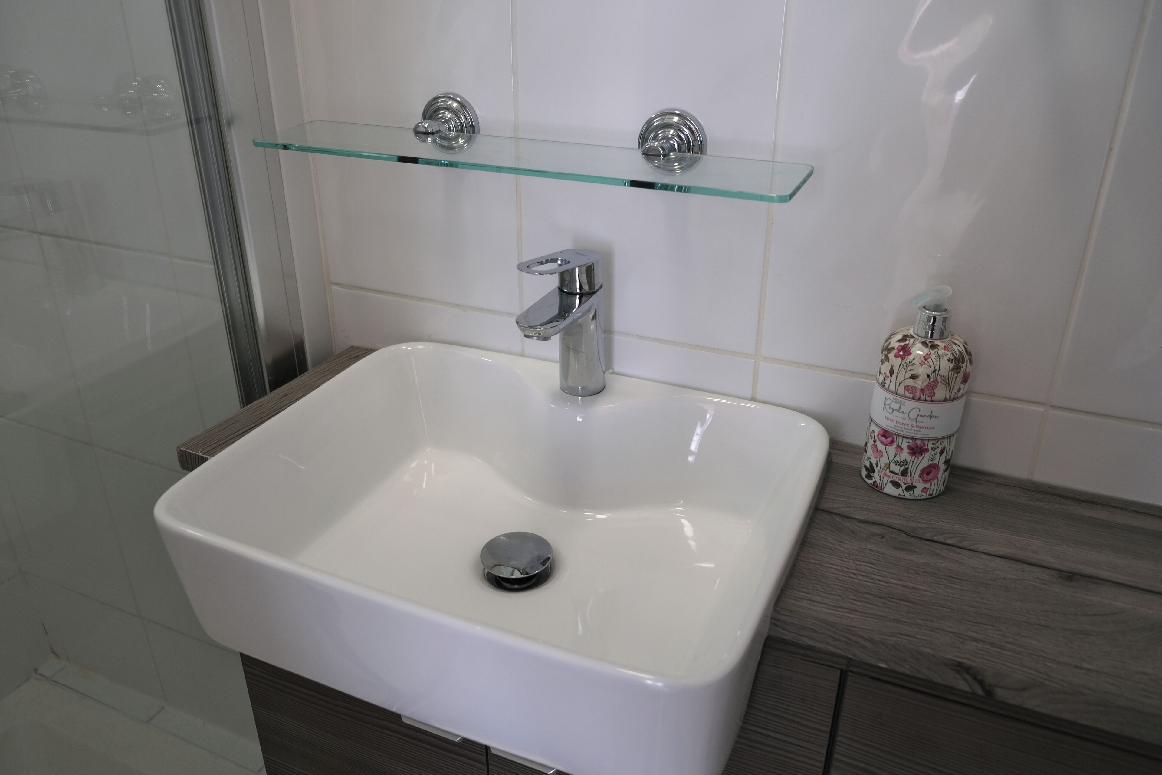 Une salle de bain avec un lavabo blanc, un robinet chromé et une étagère en verre au-dessus.