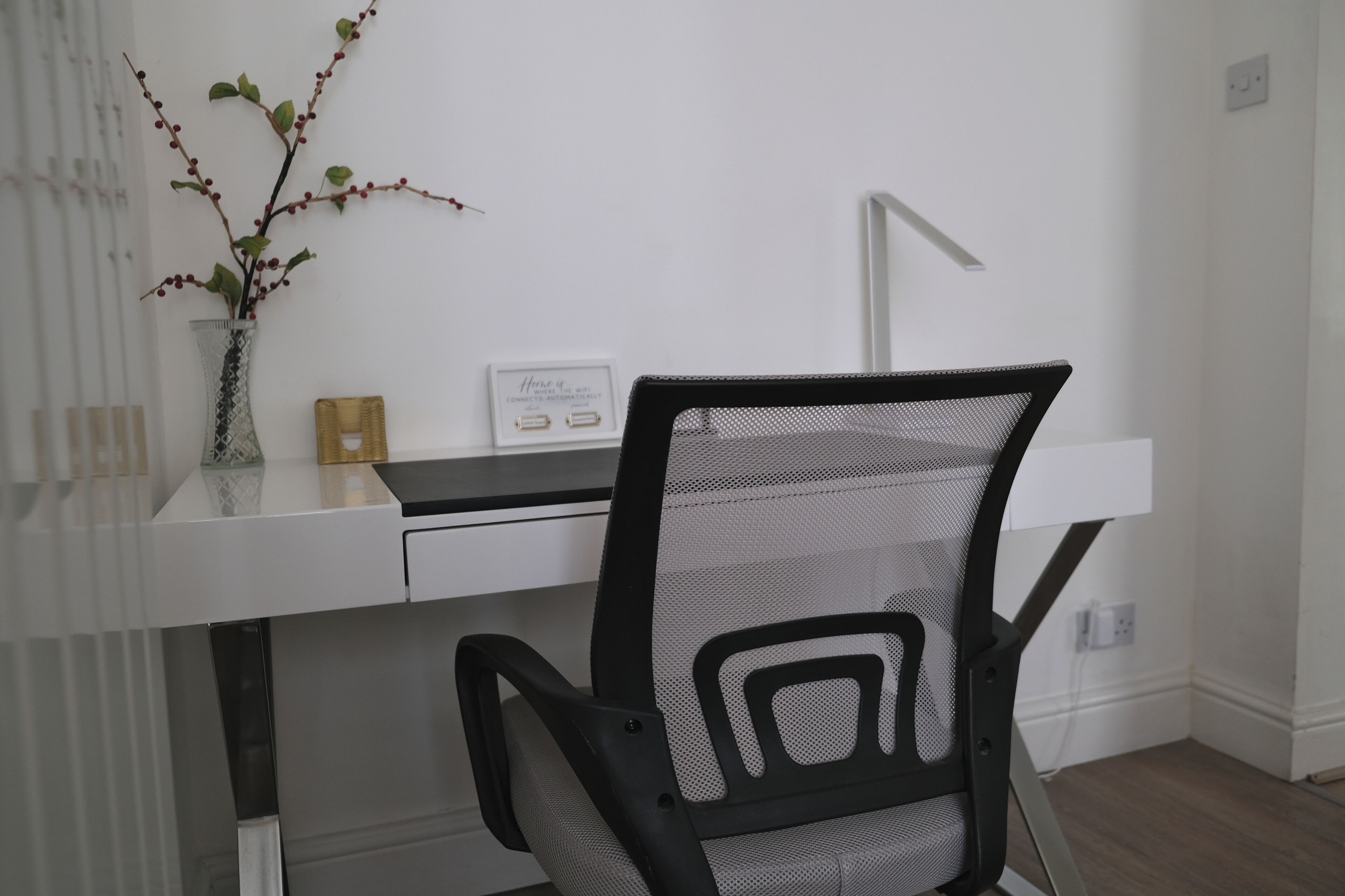 Un bureau moderne avec une chaise de bureau à dossier en maille, un vase orné de branches et un panneau décoratif encadré accroché au mur.
