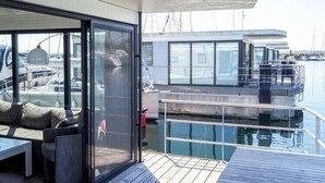 Una terraza moderna con puerta de vidrio que da a un área de descanso, con vista a un puerto deportivo lleno de barcos.