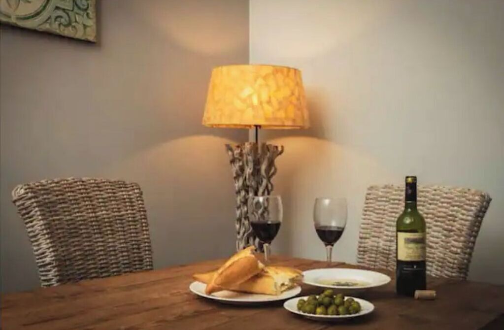 A dining table with a lamp, two chairs, a bottle of wine, a glass of wine, a plate of olives, and a plate with a sandwich.