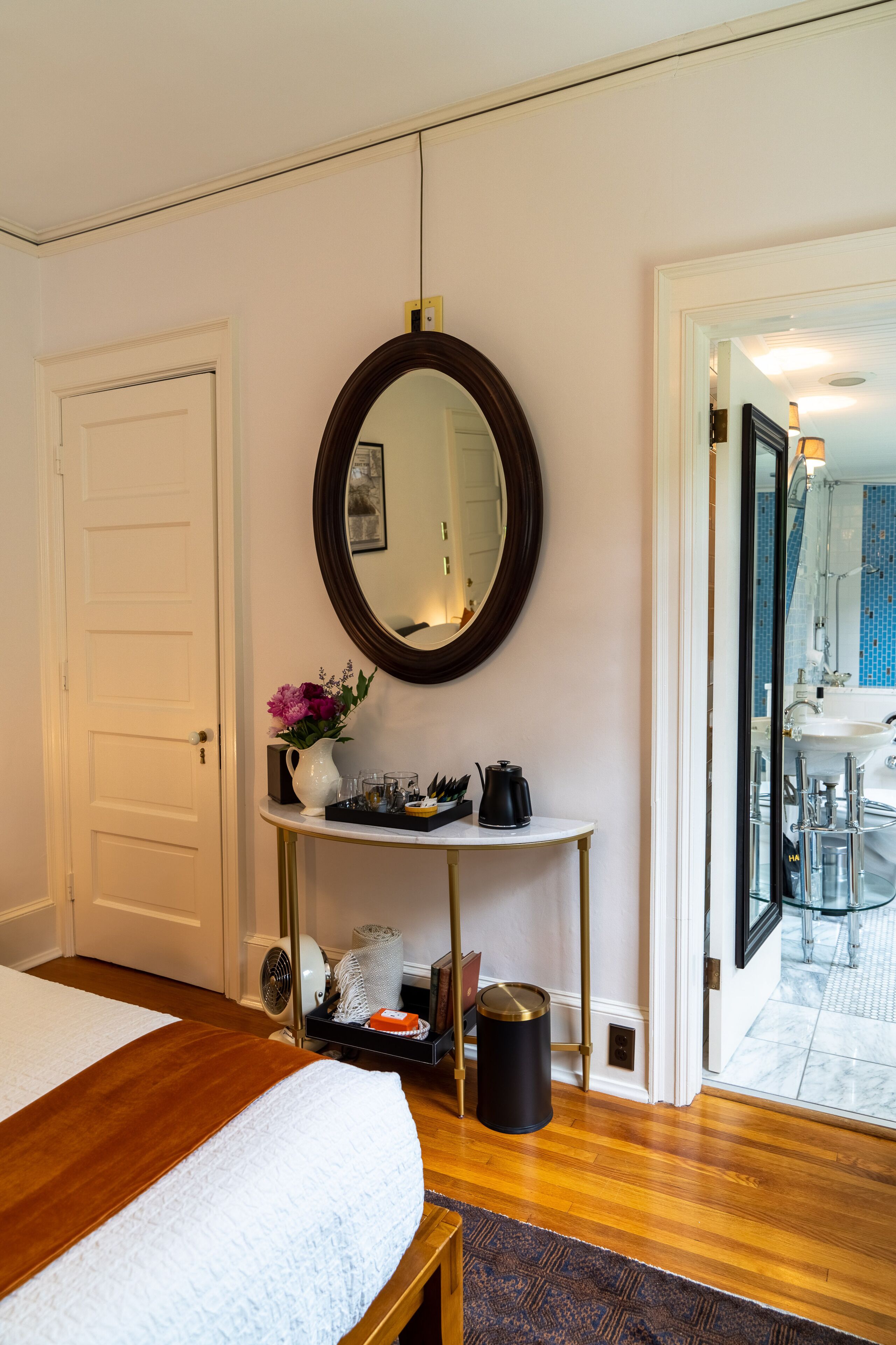 A hotel room with a bed, a bedside table with a vase of flowers, a mirror, and a view of the bathroom through an open door.