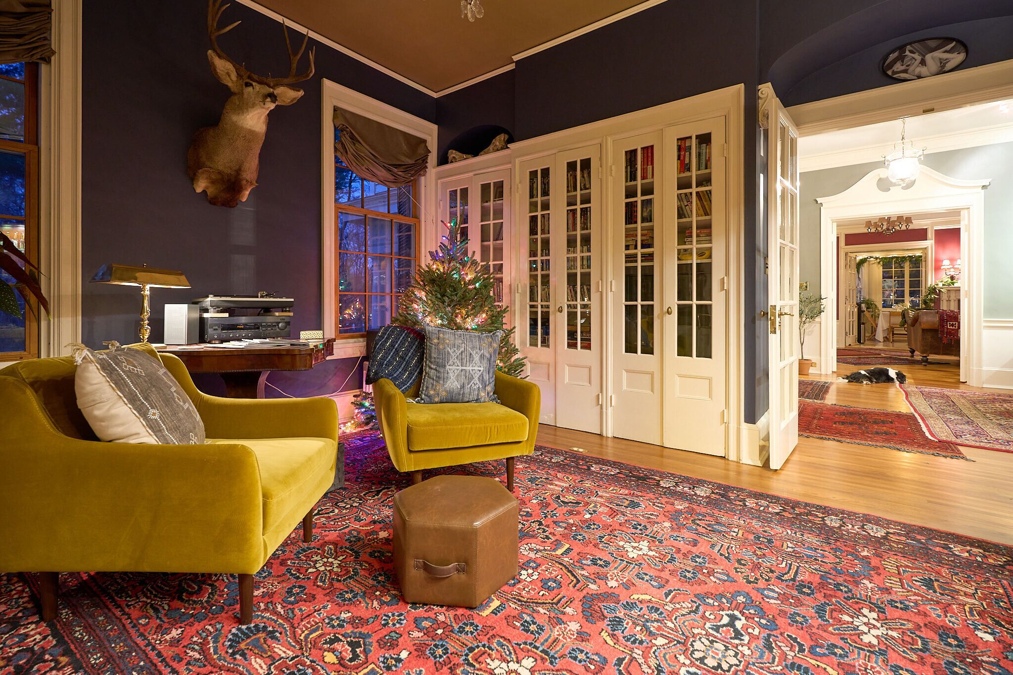 Un salon avec un fauteuil jaune, un petit pouf, un sapin de Noël et une tête de cerf accrochée au mur.