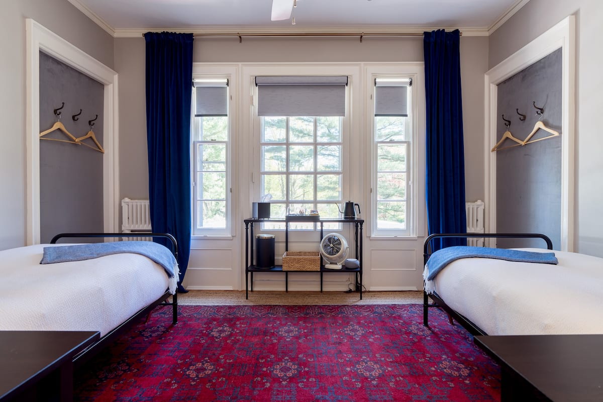 Une chambre avec deux lits, une petite table avec un ventilateur et une fenêtre avec des rideaux.