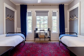 Ein Zimmer mit zwei Betten, einem kleinen Tisch mit Ventilator und einem Fenster mit Vorhängen.