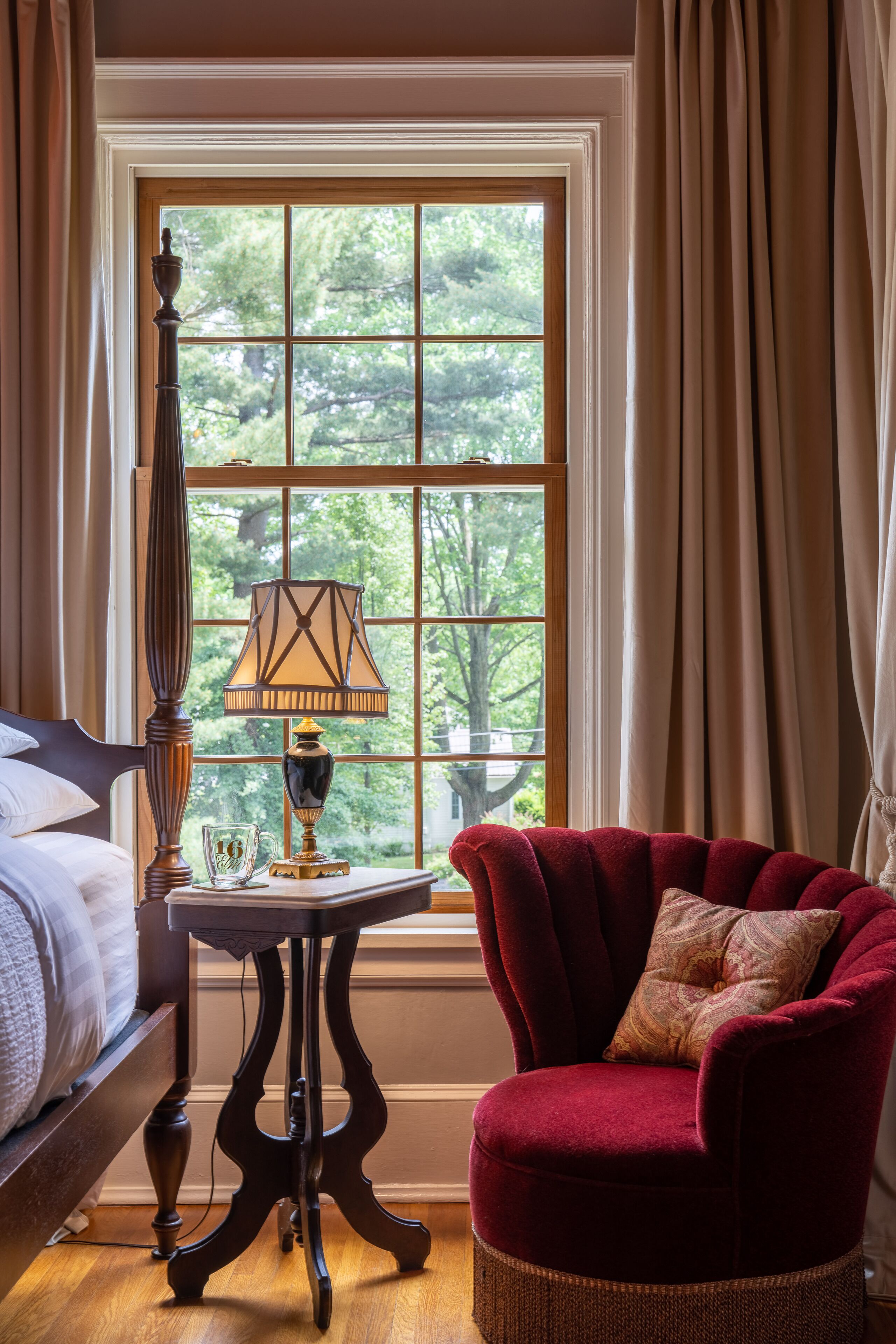 A room with a bed, a red armchair, a table with a lamp, and a window with curtains.