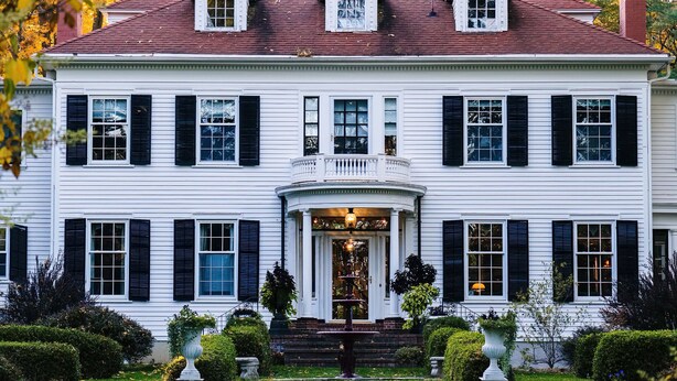Un grand bâtiment blanc avec des volets noirs, une entrée centrale avec des marches et un petit balcon au-dessus de l’entrée.