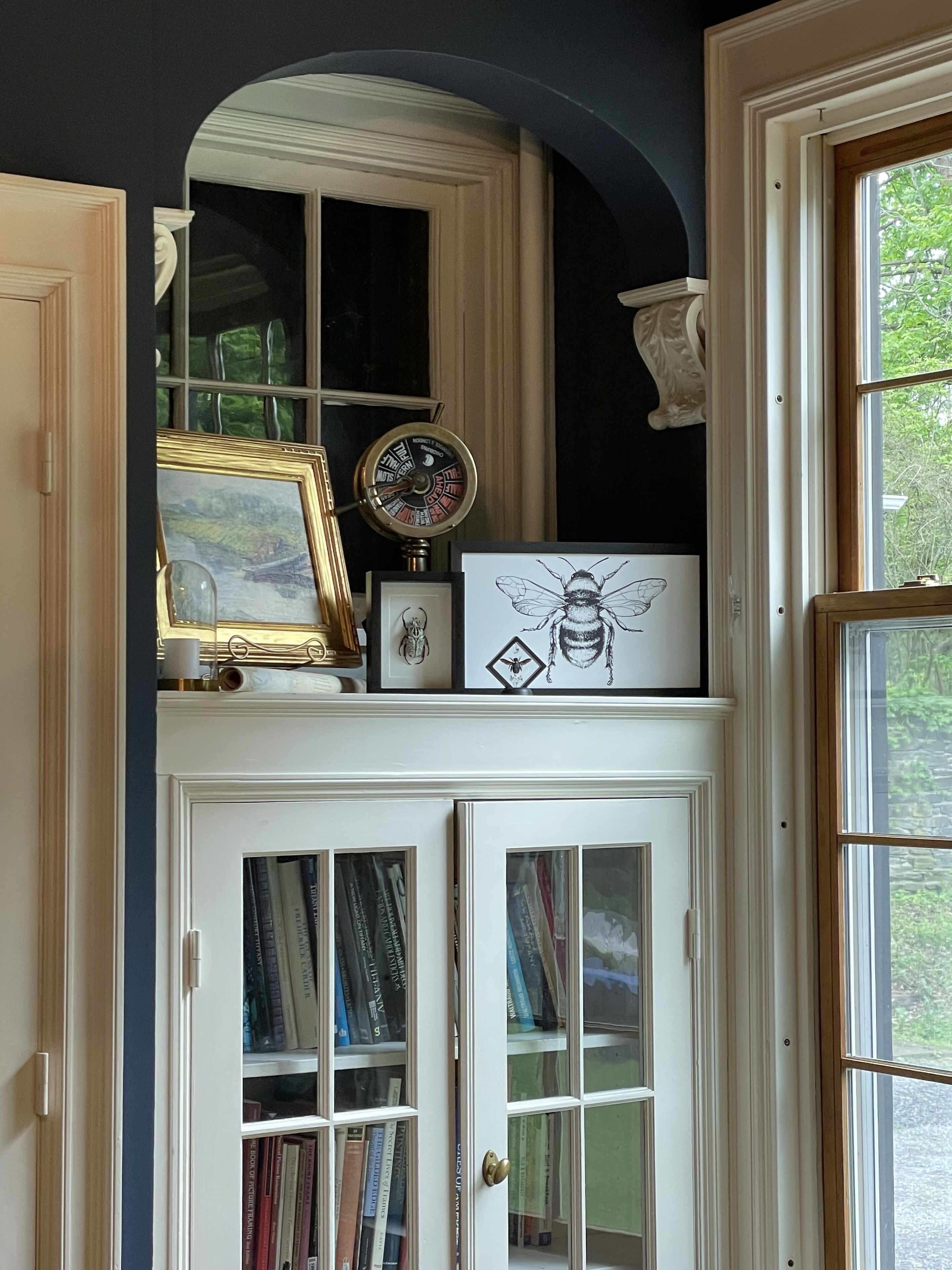 A room with a white door, a bookshelf with books, a framed picture, a clock, and a window with a view of greenery.