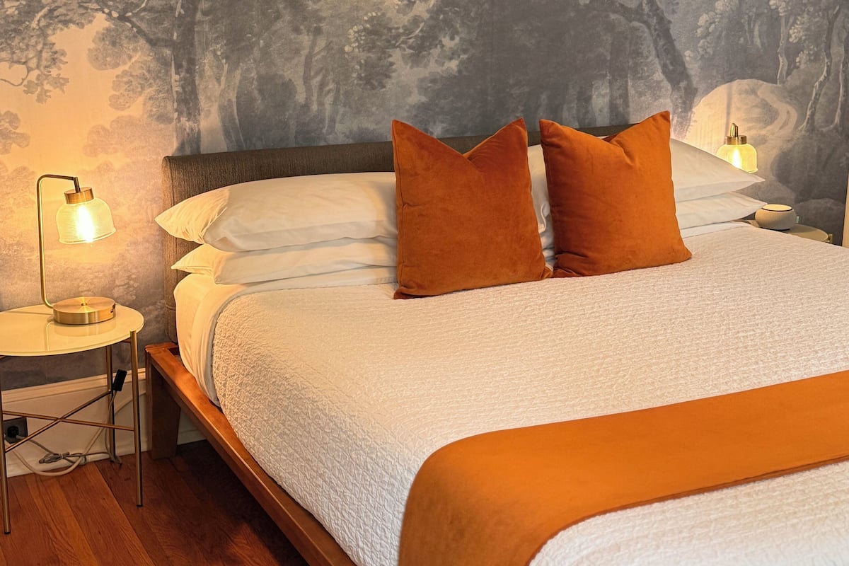 Une chambre à coucher avec un lit, deux tables de chevet, une lampe et un mur orné d’une fresque représentant un arbre.