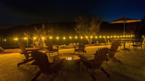 Un espace extérieur aménagé avec des chaises en bois et un foyer, entouré d’une petite clôture et illuminé de guirlandes lumineuses.