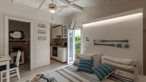 Een ruime woonkamer met een bank, een gestreept vloerkleed en een eettafel. Achtergrond is een keuken met witte kasten en een raam.