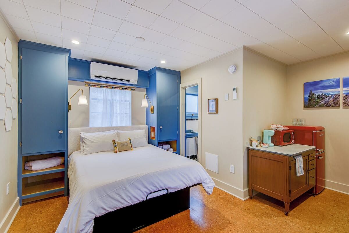 A bedroom with a bed, a wardrobe, an air conditioning unit, a small kitchenette, and a bathroom.