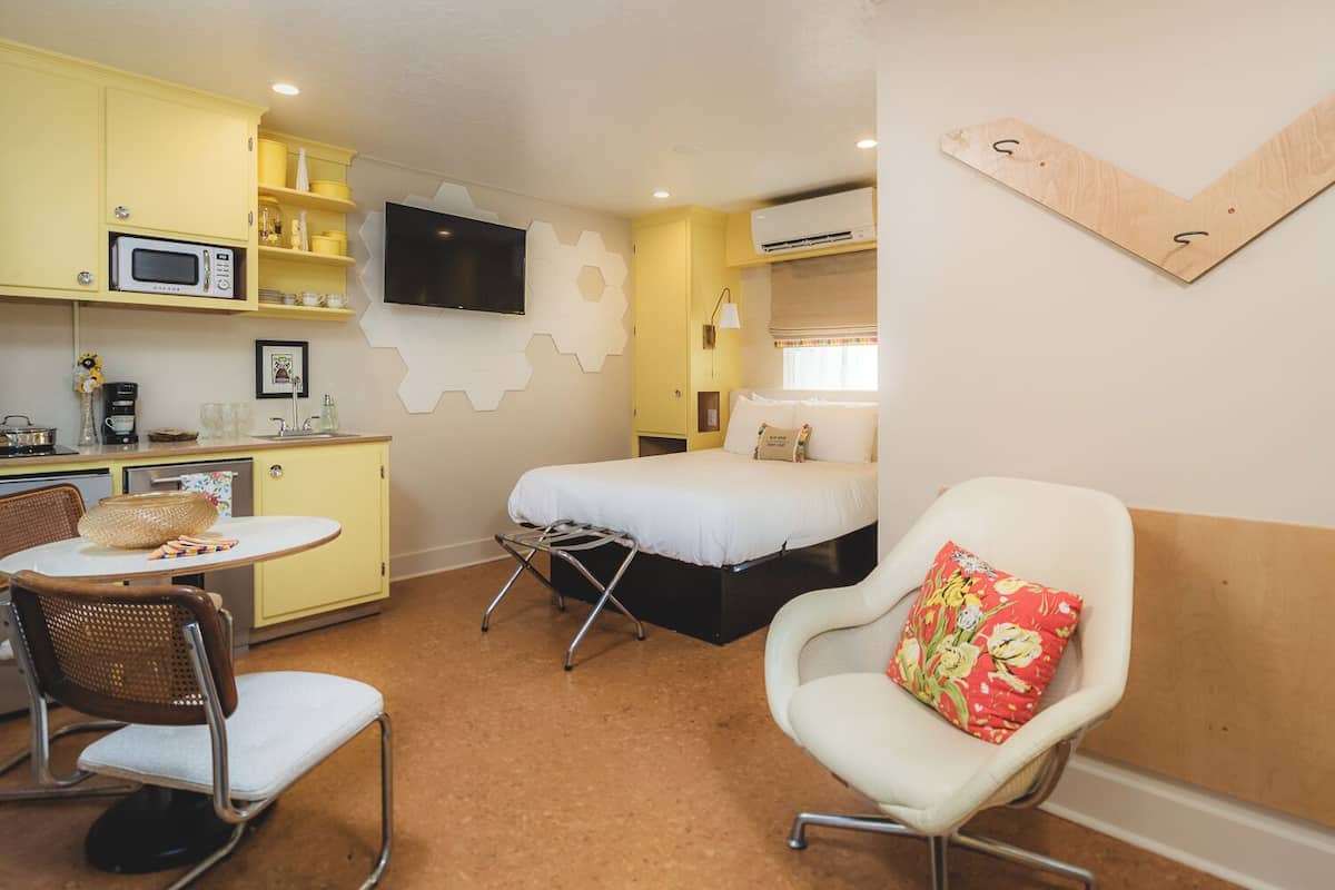 A compact hotel room with a bed, a small dining area with a table and chairs, a microwave, a TV, and an air conditioning unit.