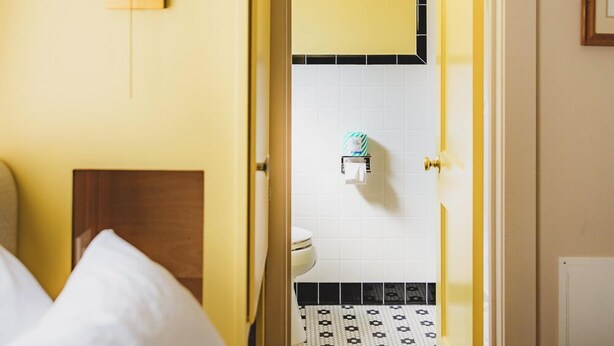 A hotel room with a bed, a nightstand, a wall-mounted lamp, a bathroom with a toilet and sink, and a yellow door. - Blue Ridge Tourist Court (Boone)