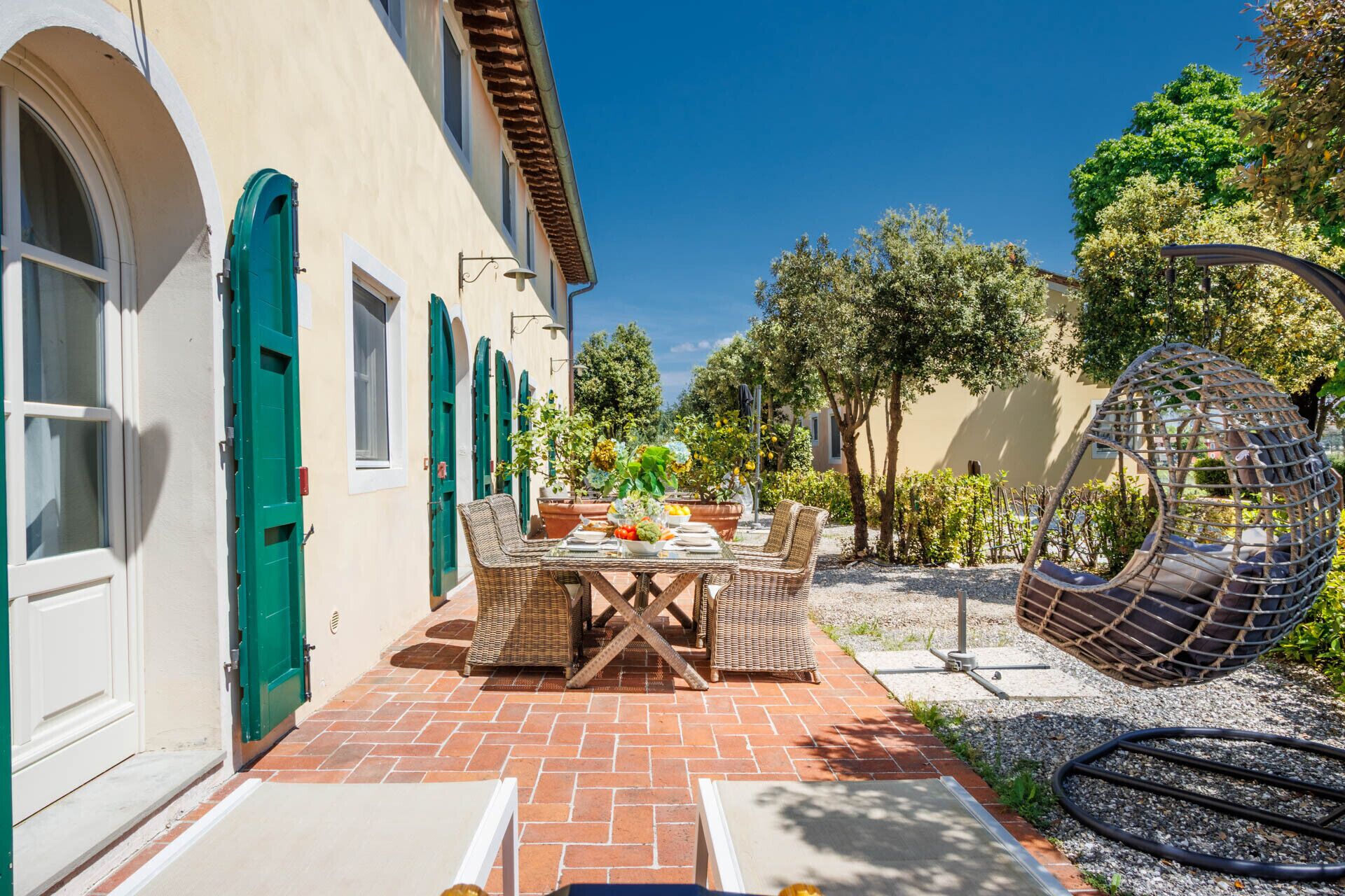 A patio with a table set for dining, surrounded by a building with green shutters and a hanging chair.