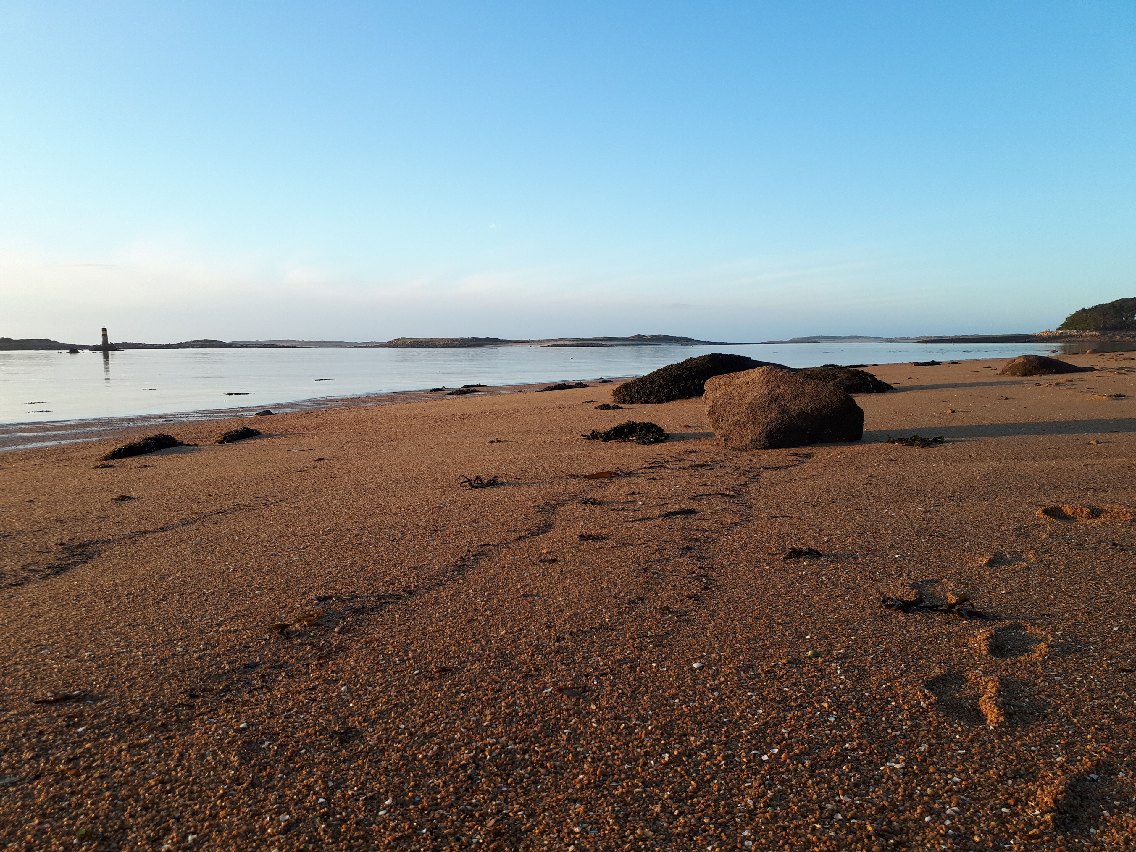 Ein kiesiger Strand mit großen Felsen und einem Leuchtturm in der Ferne.