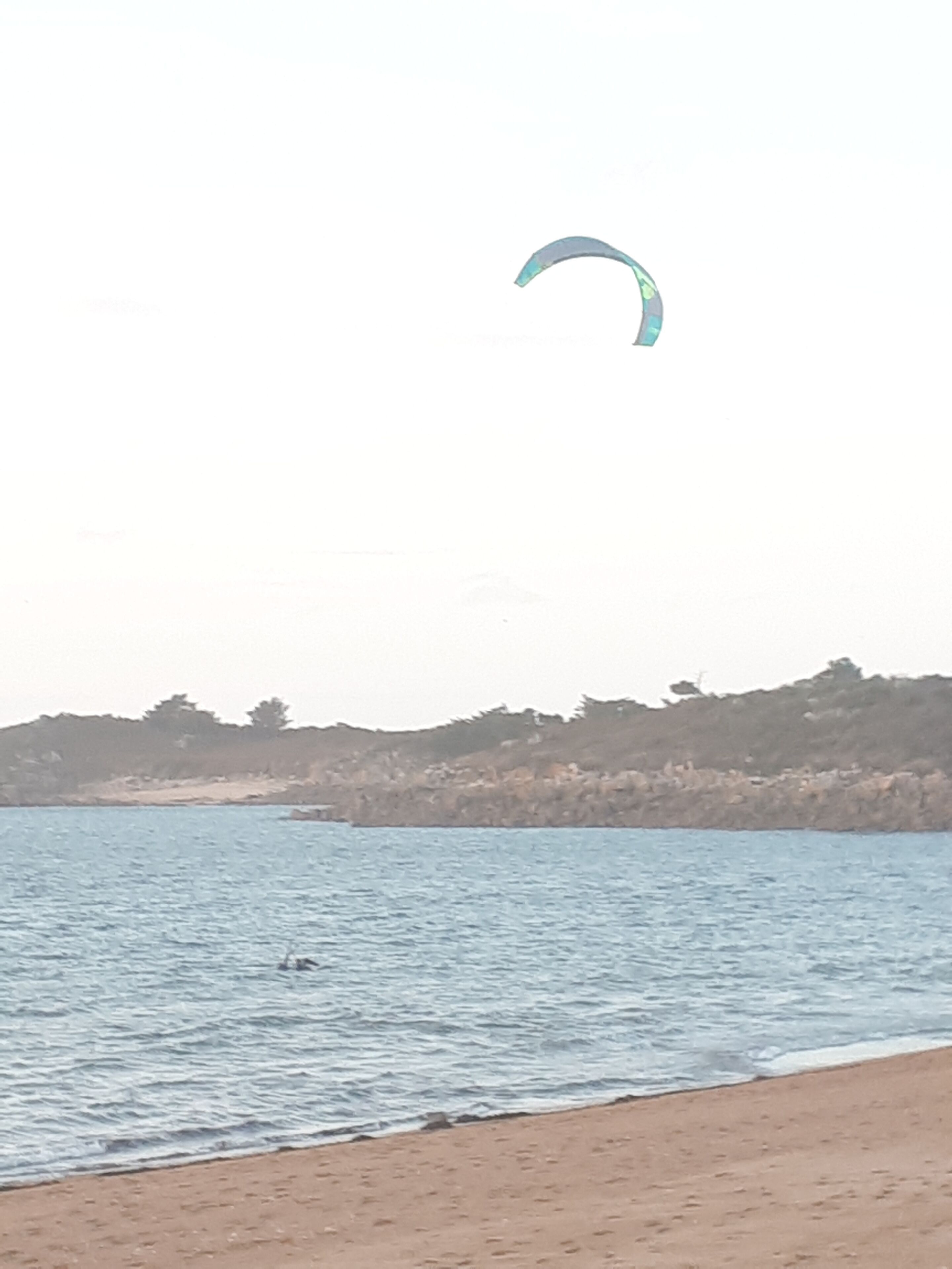 Una playa con un paracaídas en el cielo, agua tranquila y una costa distante con árboles.