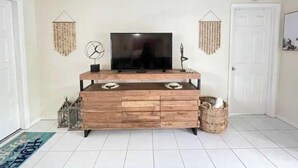 A living room with a wooden entertainment unit, a flat-screen TV, decorative wall hangings, and a white door. - PGA 3/2 w Backyard close to everything beach mall (West Palm Beach)