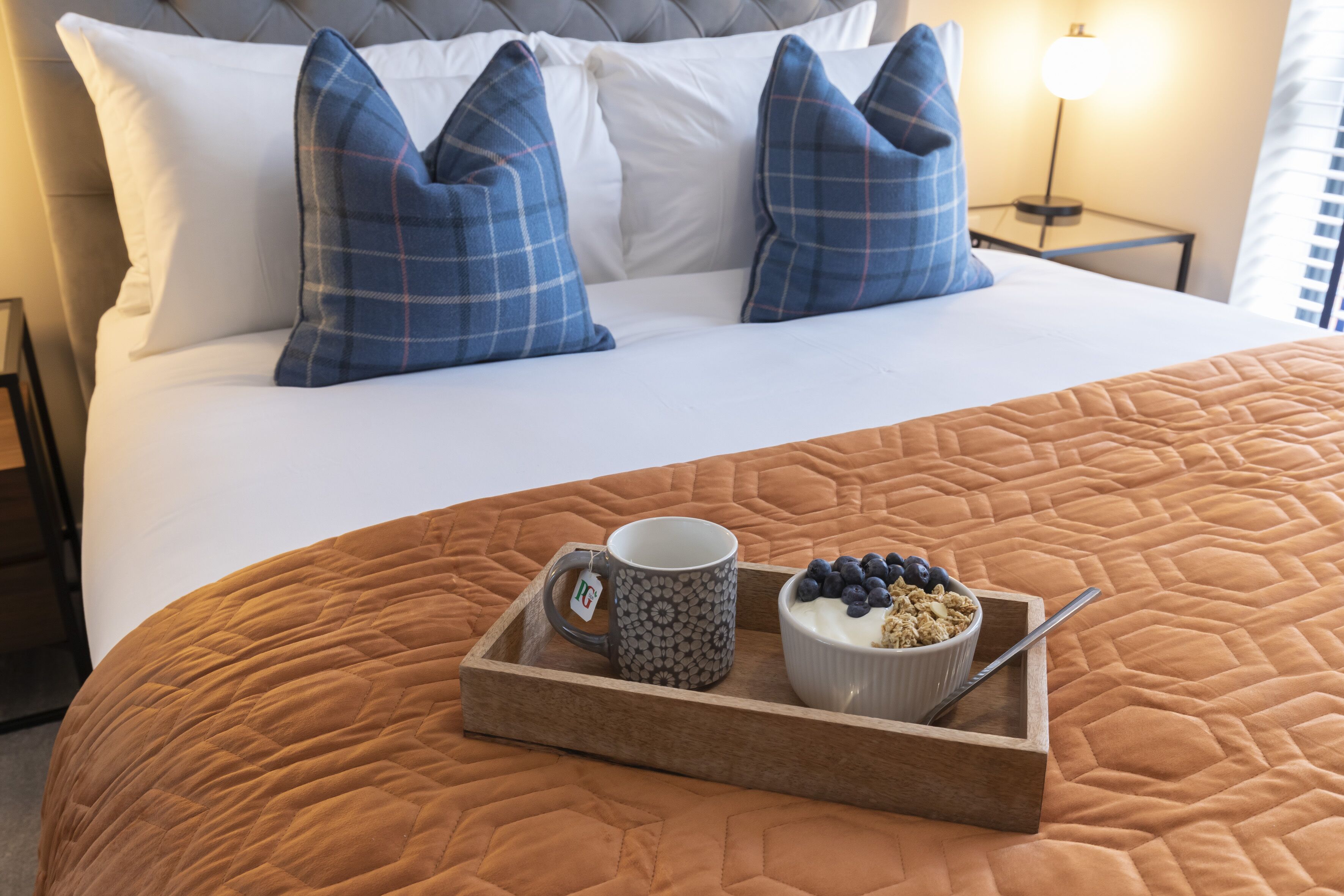 Un lit bien fait, recouvert d’une couverture marron, avec deux coussins à carreaux bleus, un plateau sur lequel se trouvent une tasse, un bol de yaourt aux myrtilles et une cuillère.