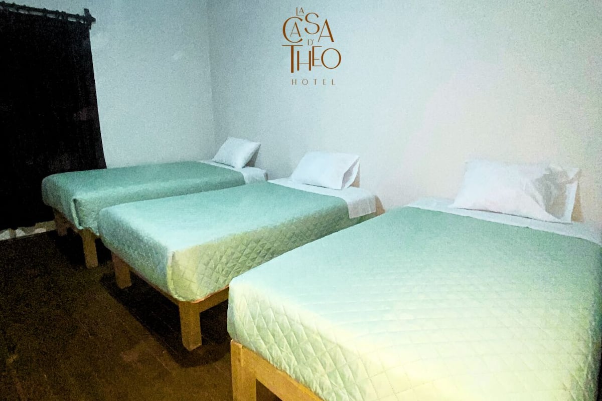 A hotel room with three single beds, each with a quilted mattress and white pillows. The room has a wooden floor and white walls.