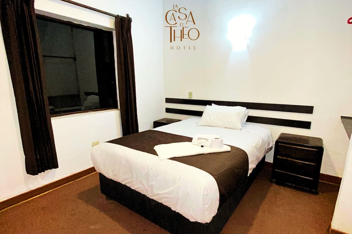A hotel room with a large bed, a bedside table, a nightstand, and a window with curtains.