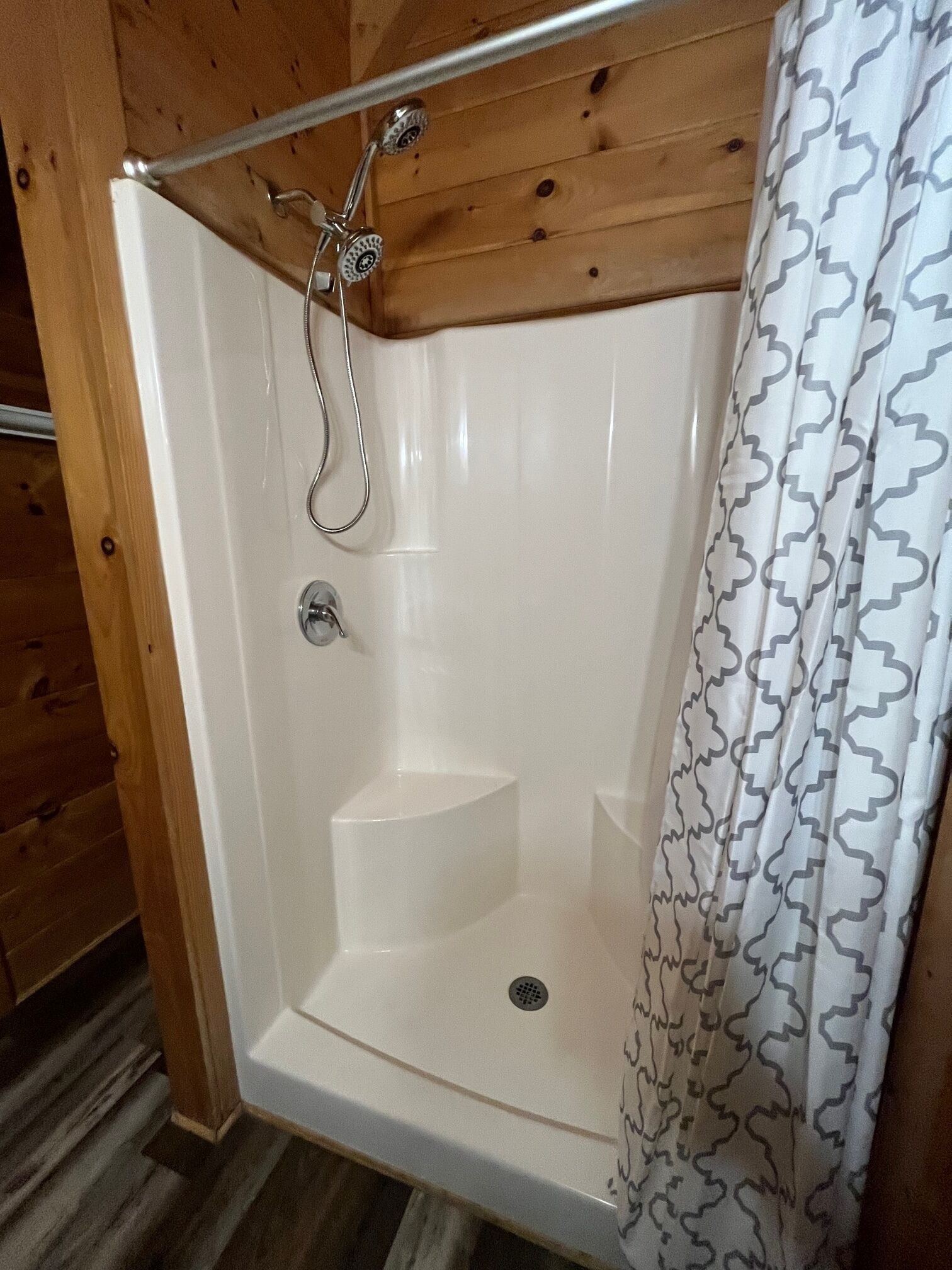 A shower with a metal shower head and a drain, a wooden ceiling, and a patterned shower curtain.