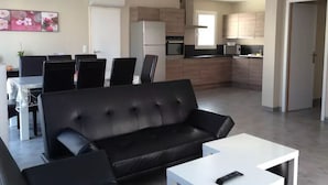 Un saló modern amb un sofà de pell negra, una taula de centre blanca i una zona de menjador amb una taula i cadires. Es veu una cuina amb armaris de fusta i electrodomèstics al fons.