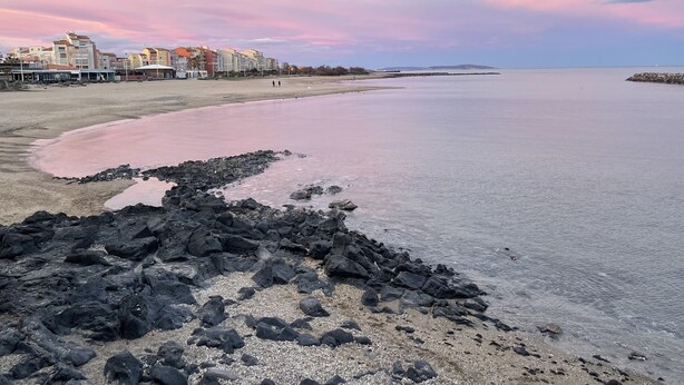 Una platja amb primer terme rocos, un mar tranquil i edificis al fons.