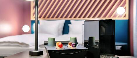 Une chambre d’hôtel bien aménagée, avec un lit moderne, une table de chevet dotée d’une lampe, un haut-parleur et un plateau avec des tasses et une bouteille.
