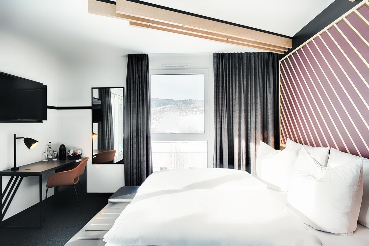 Una habitación de hotel moderna con una cama grande, un escritorio con silla, un televisor de pantalla plana y una ventana con cortinas.