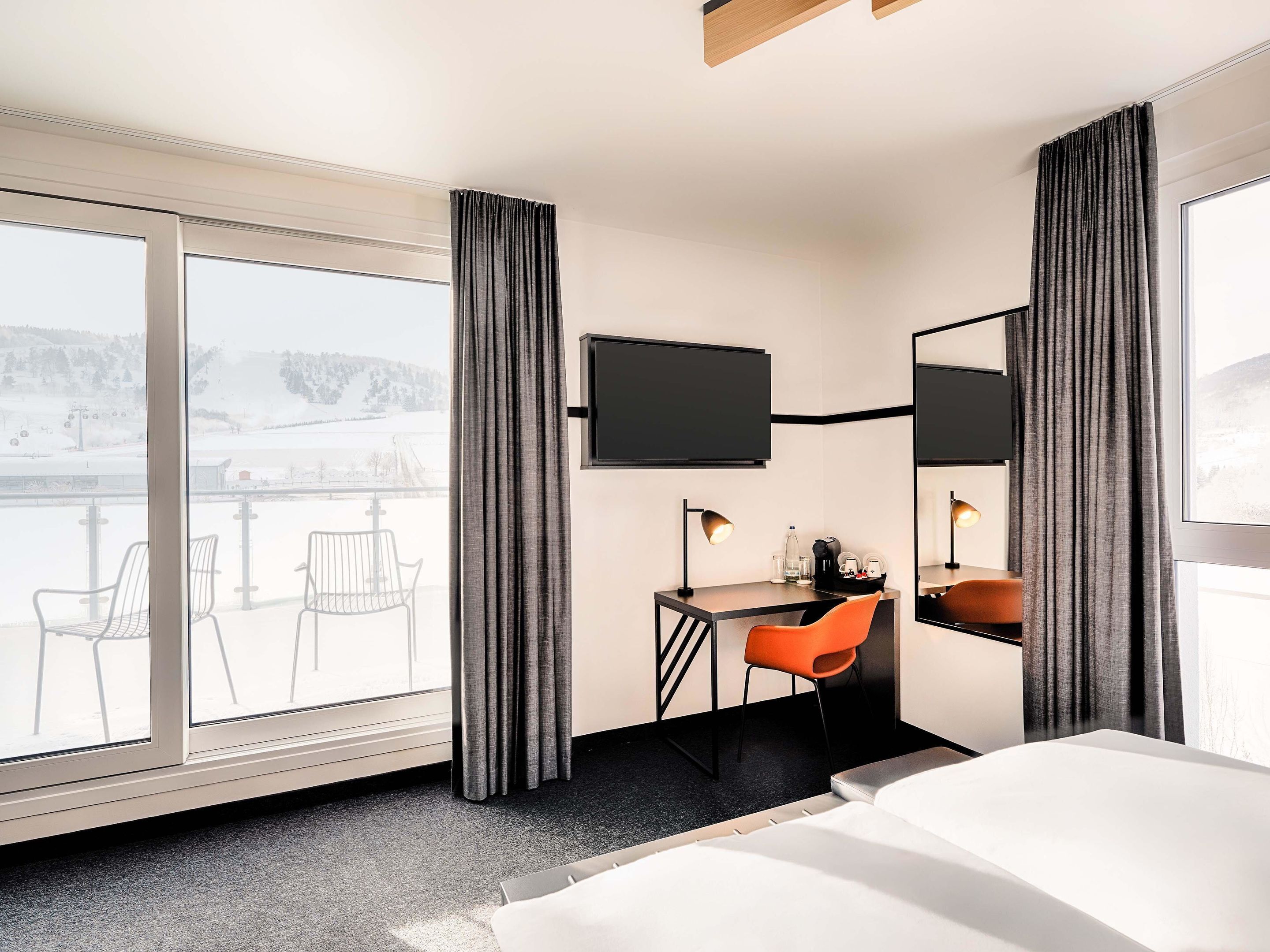 Ein Hotelzimmer mit einem Bett, einem Schreibtisch mit Stuhl, einem an der Wand befestigten Fernseher und einem großen Fenster mit Vorhängen.