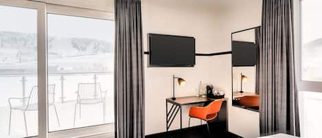 A hotel room with a bed, a desk with a chair, a TV mounted on the wall, and a large window with curtains.