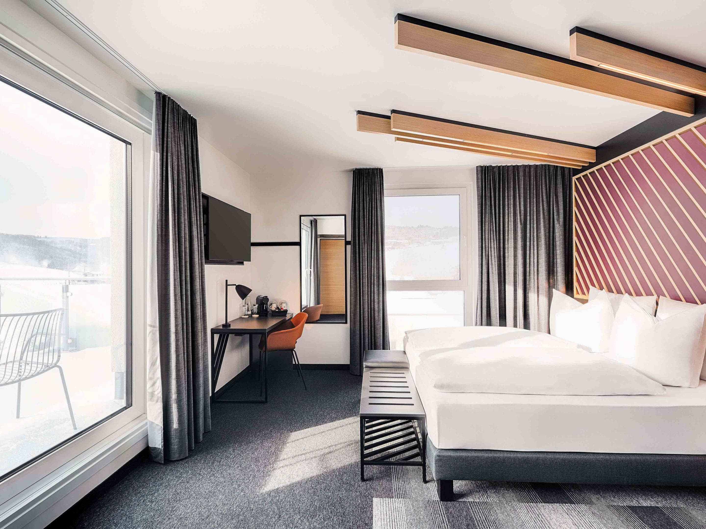 A modern hotel room with a large bed, a desk, and a chair. There is a TV mounted on the wall, and the room has large windows with curtains.
