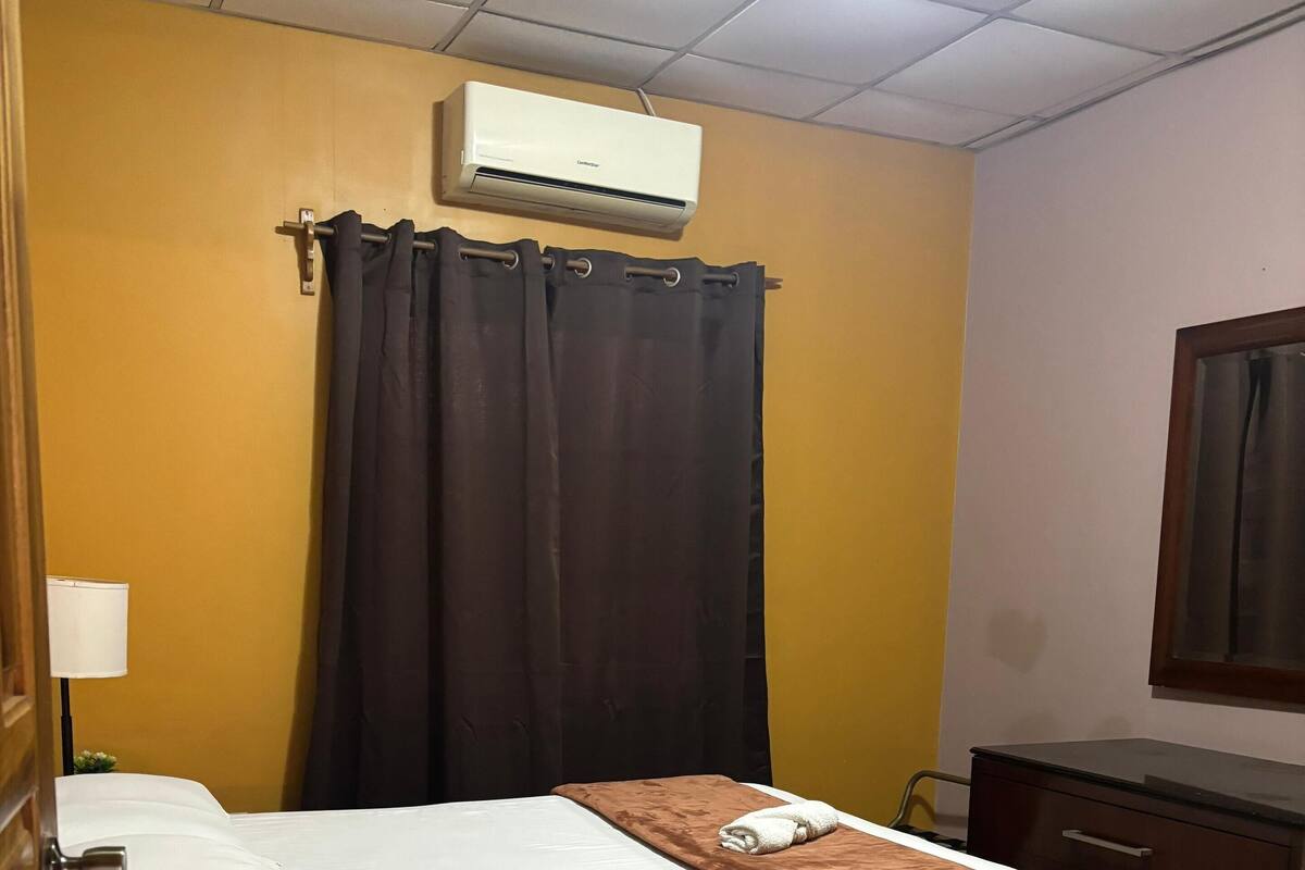 A hotel room with a bed, a bedside table, a lamp, a brown throw blanket, a curtain, and an air conditioning unit on the wall.