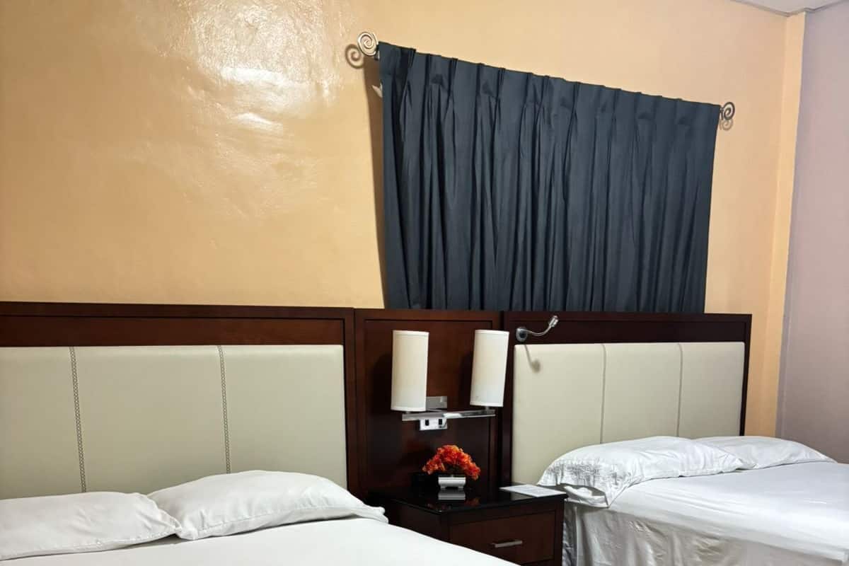 A hotel room with two beds, a nightstand with a lamp, and a window with a dark curtain.