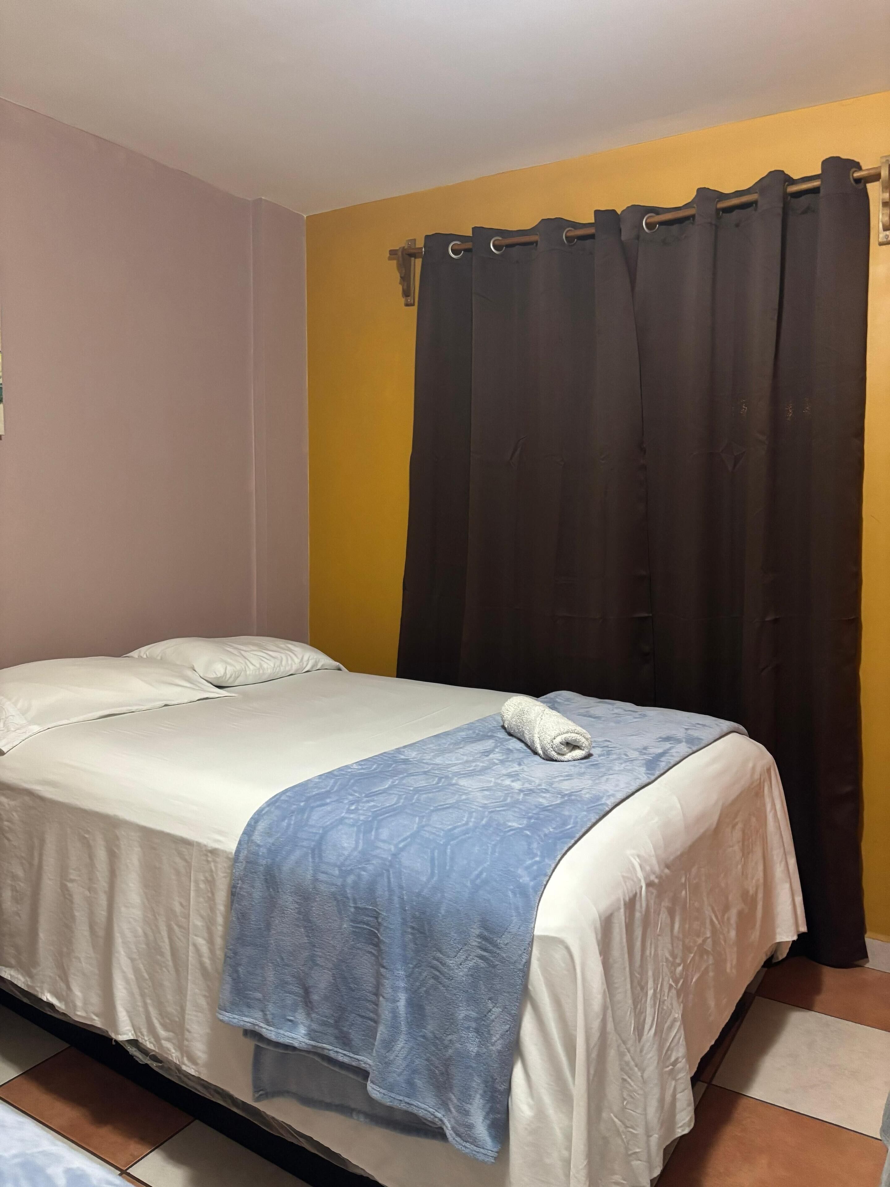 A single bed with a white and blue bedspread, a rolled towel, and a dark curtain.
