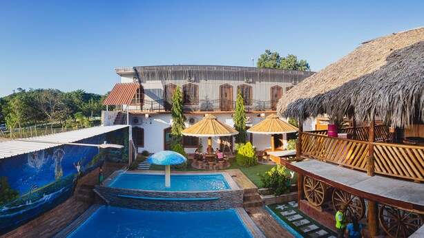 A pool area with a mural, a thatched-roof structure, and a building with arched windows. - Hotel y Restaurante Las Adas y Estrellas (Melchor de Mencos)